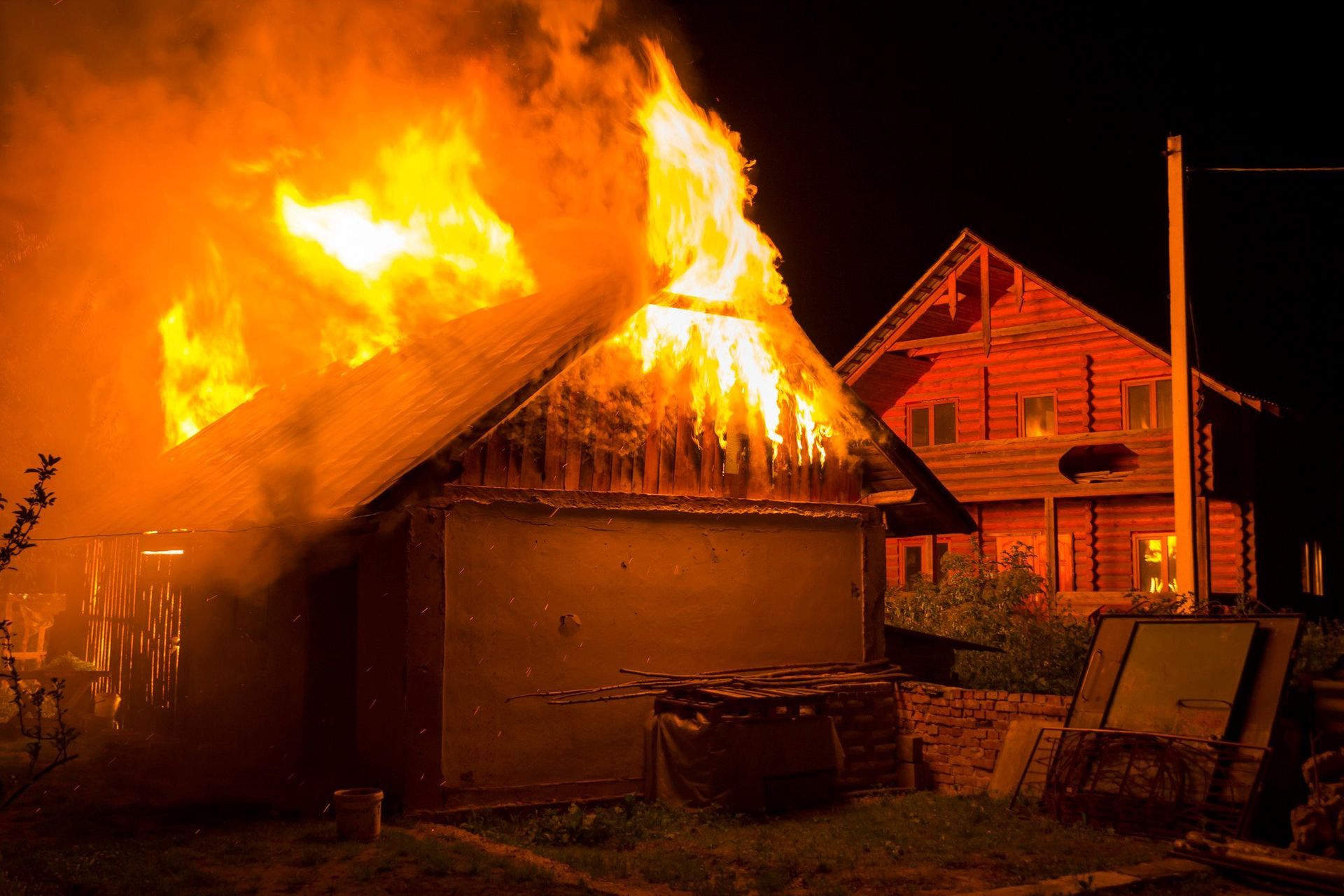 House roof engulfed in bright orange flames at night, with a nearby home in the background.