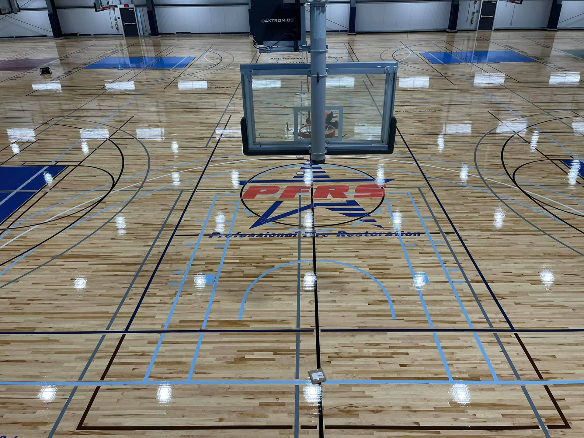 Empty indoor basketball court with a centered hoop and glossy wood floor marked with blue and black lines.