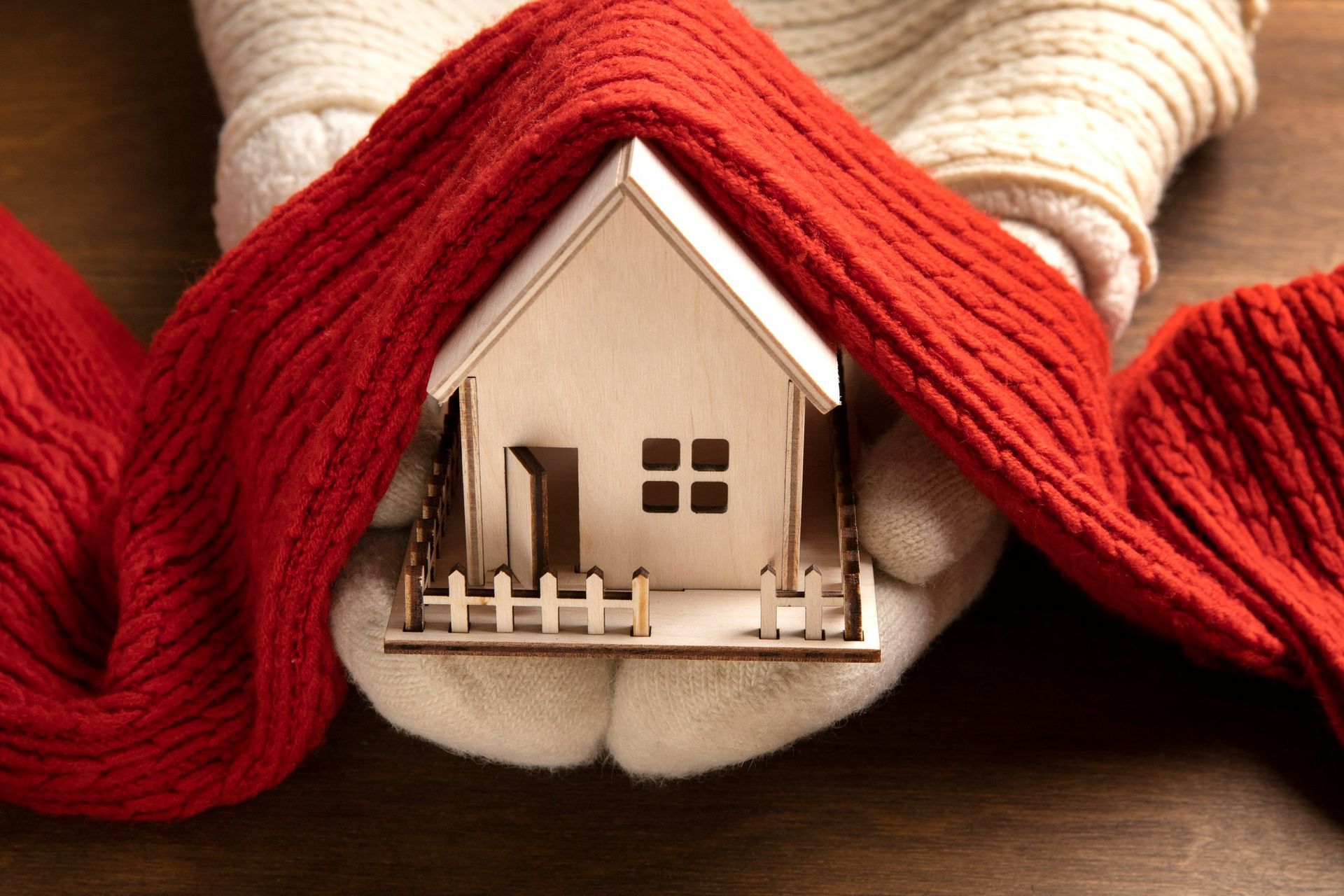 Small house model wrapped in a red scarf, held by gloved hands on a wooden surface