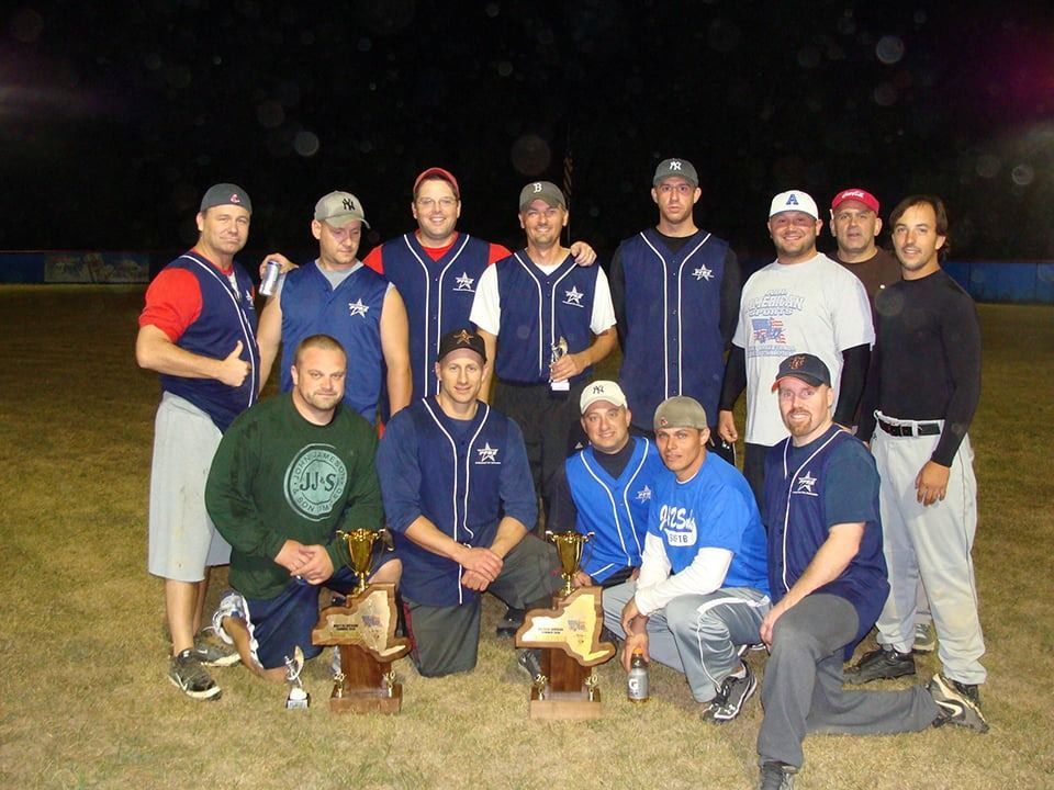 Group of people posing outdoors at night with trophies and a dog, wearing matching navy team shirts.
