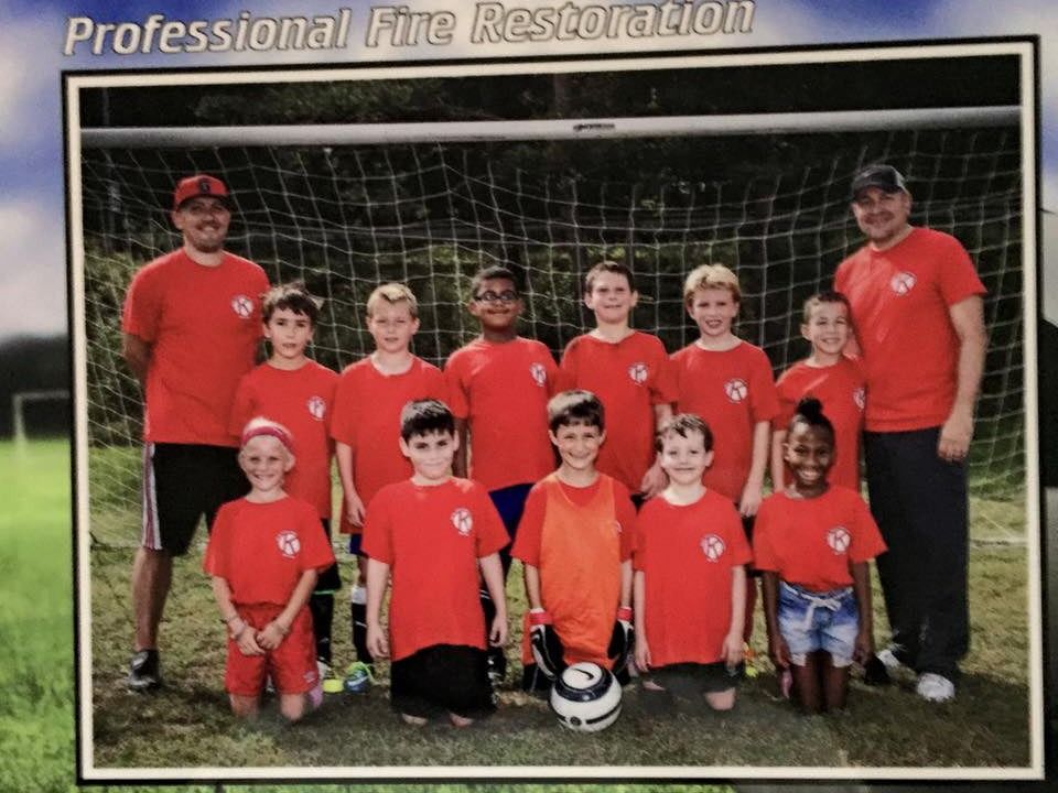 Youth soccer team posing in red jerseys with coaches in front of a goal and sponsor banner.