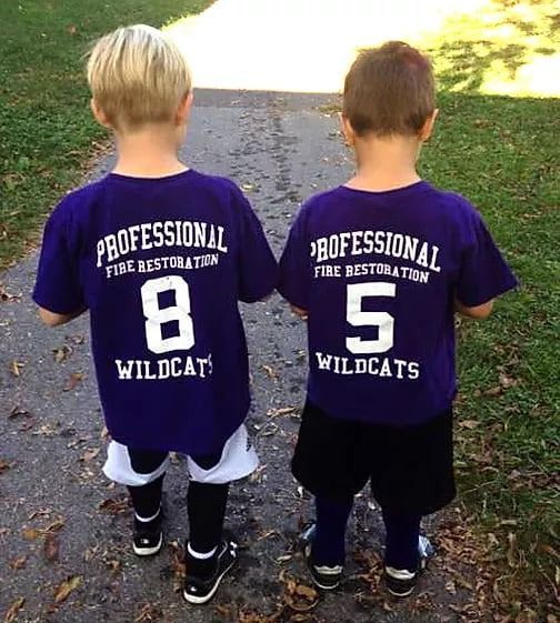 Two children from behind wearing matching purple firefighter shirts with numbers 8 and 5, standing outdoors.