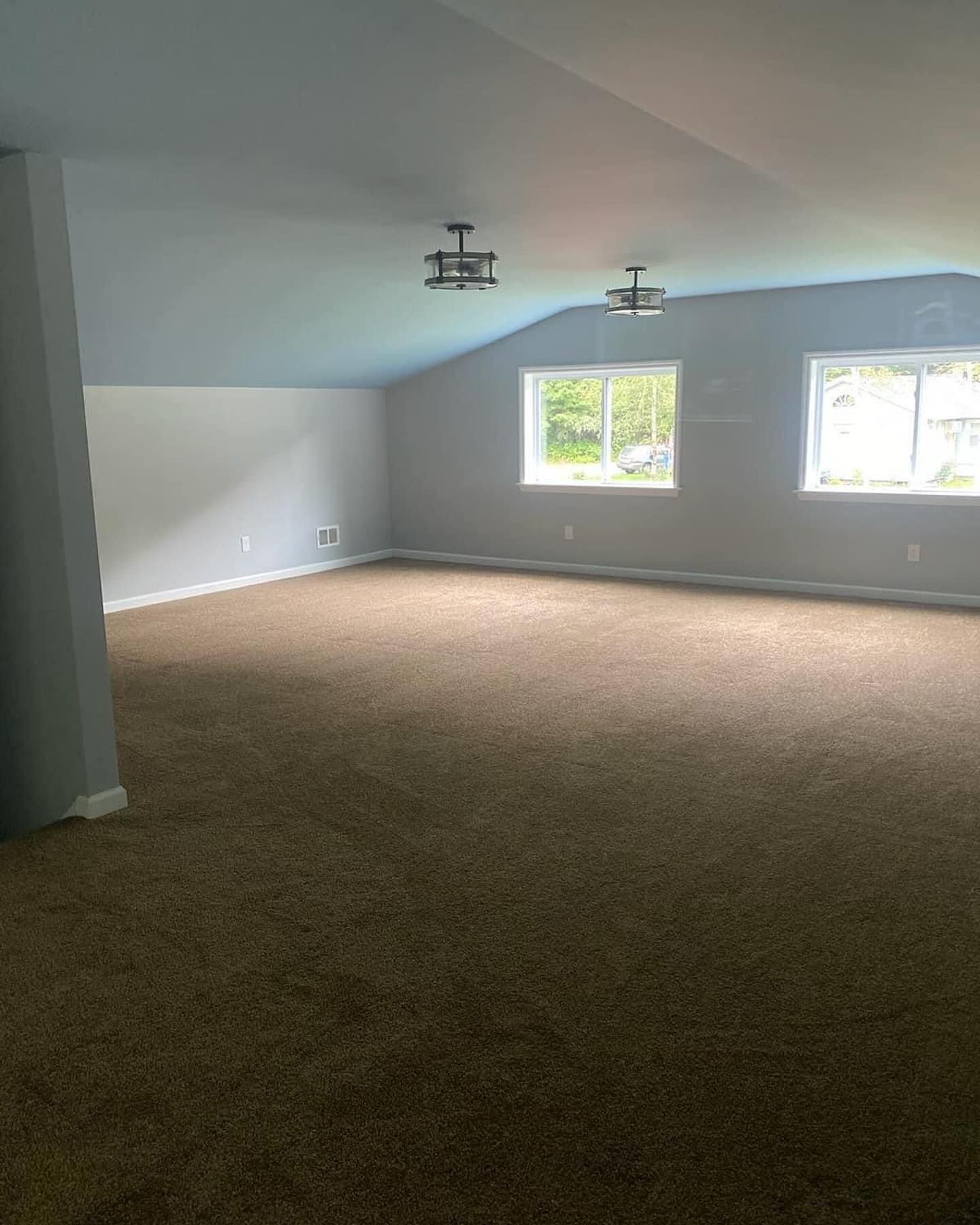 Empty carpeted room with sloped ceiling and two windows letting in daylight.