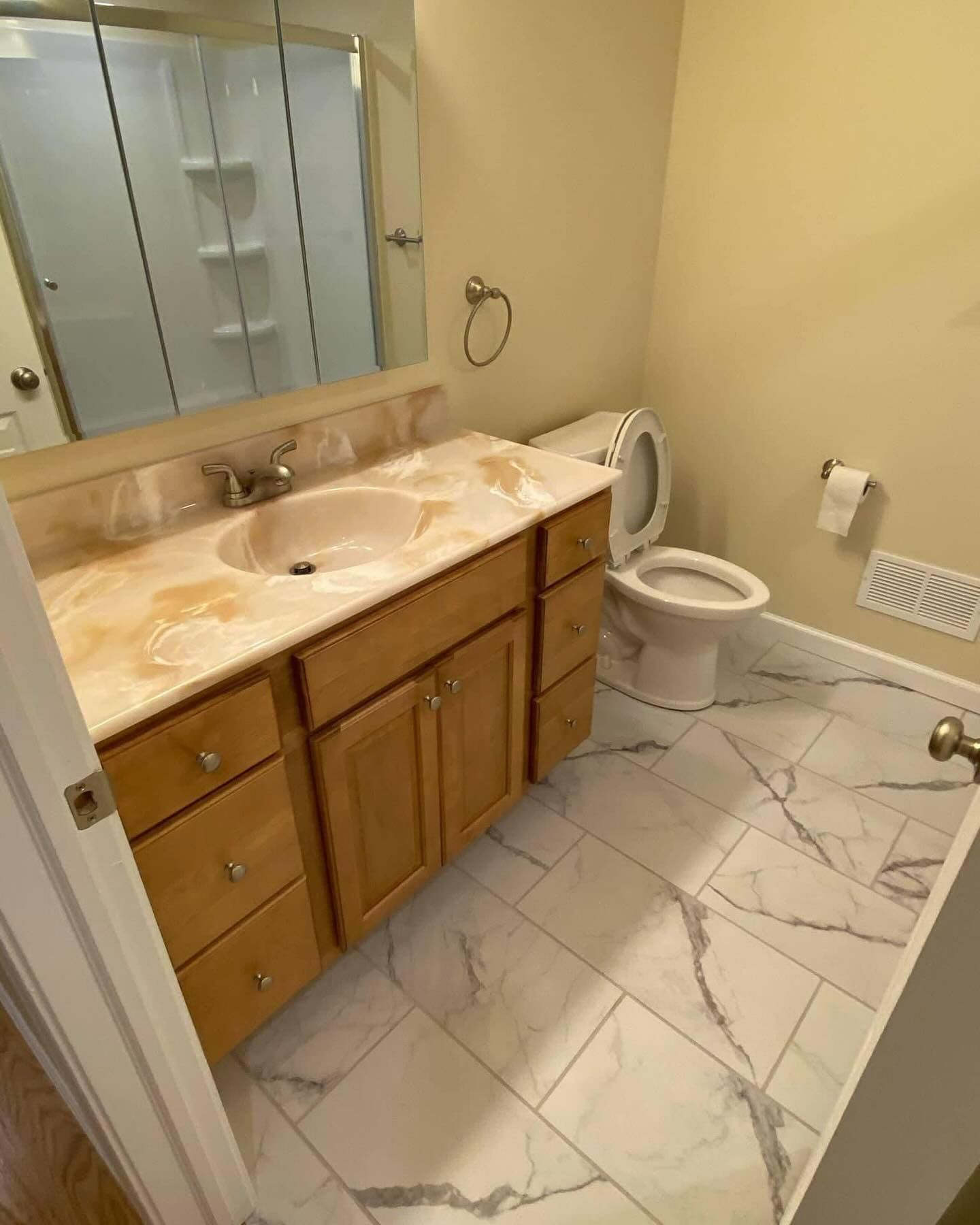 Small bathroom with wood vanity, marble-pattern floor, toilet, sink, mirror, and shower stall.