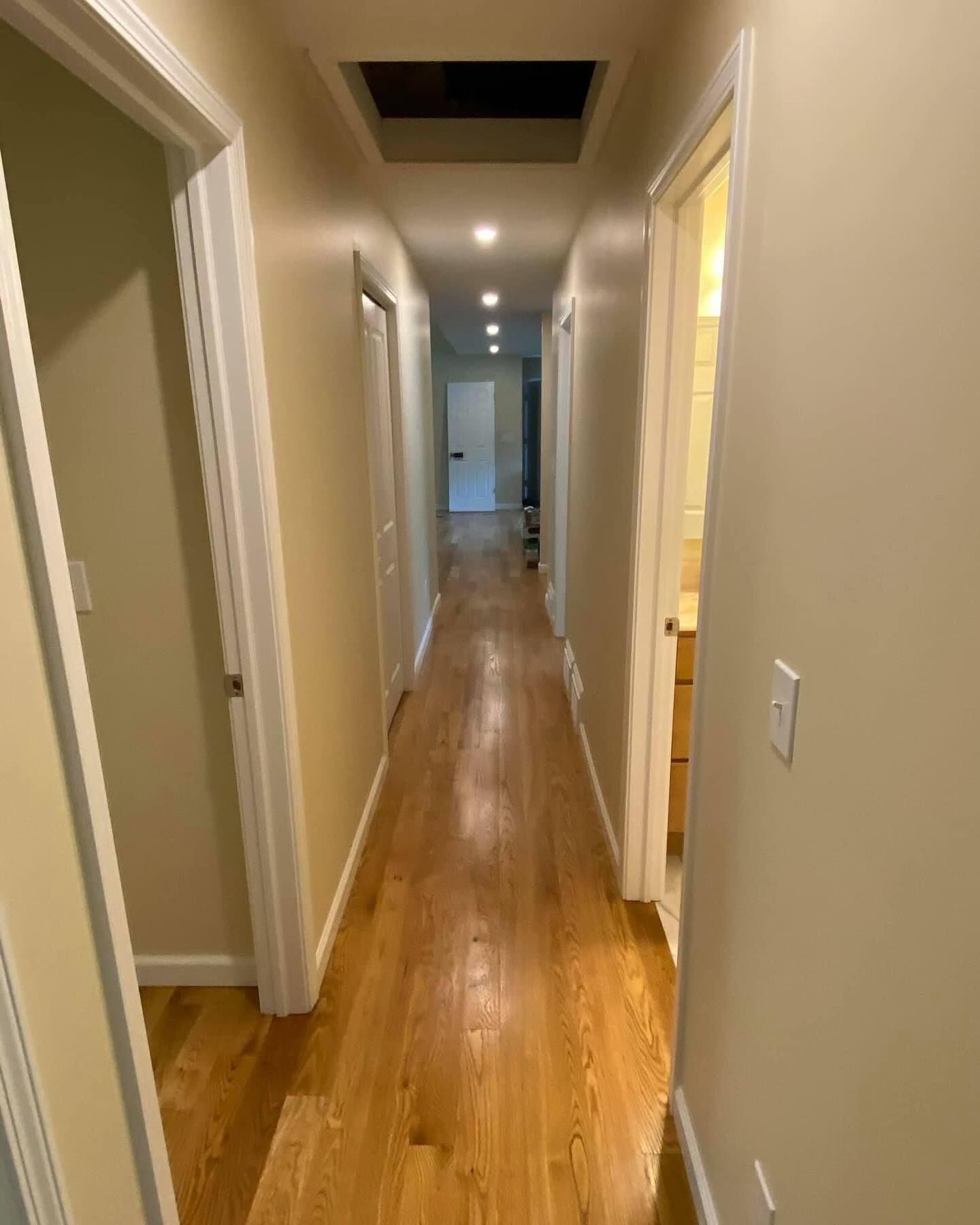 Narrow beige hallway with hardwood floors, white doors, and ceiling lights leading to a distant room