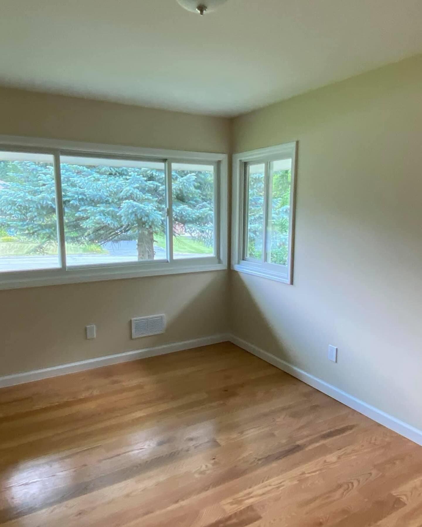Empty beige room with hardwood floors and large windows looking out to green trees