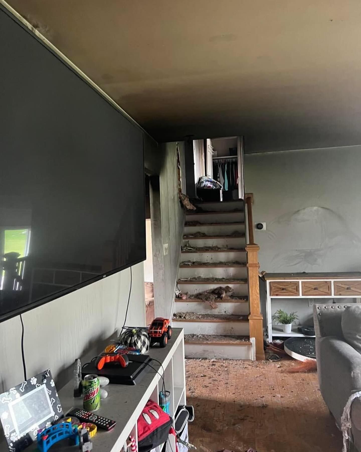 Dim living room with stairs, TV on left wall, and cluttered shelves in a partially renovated space