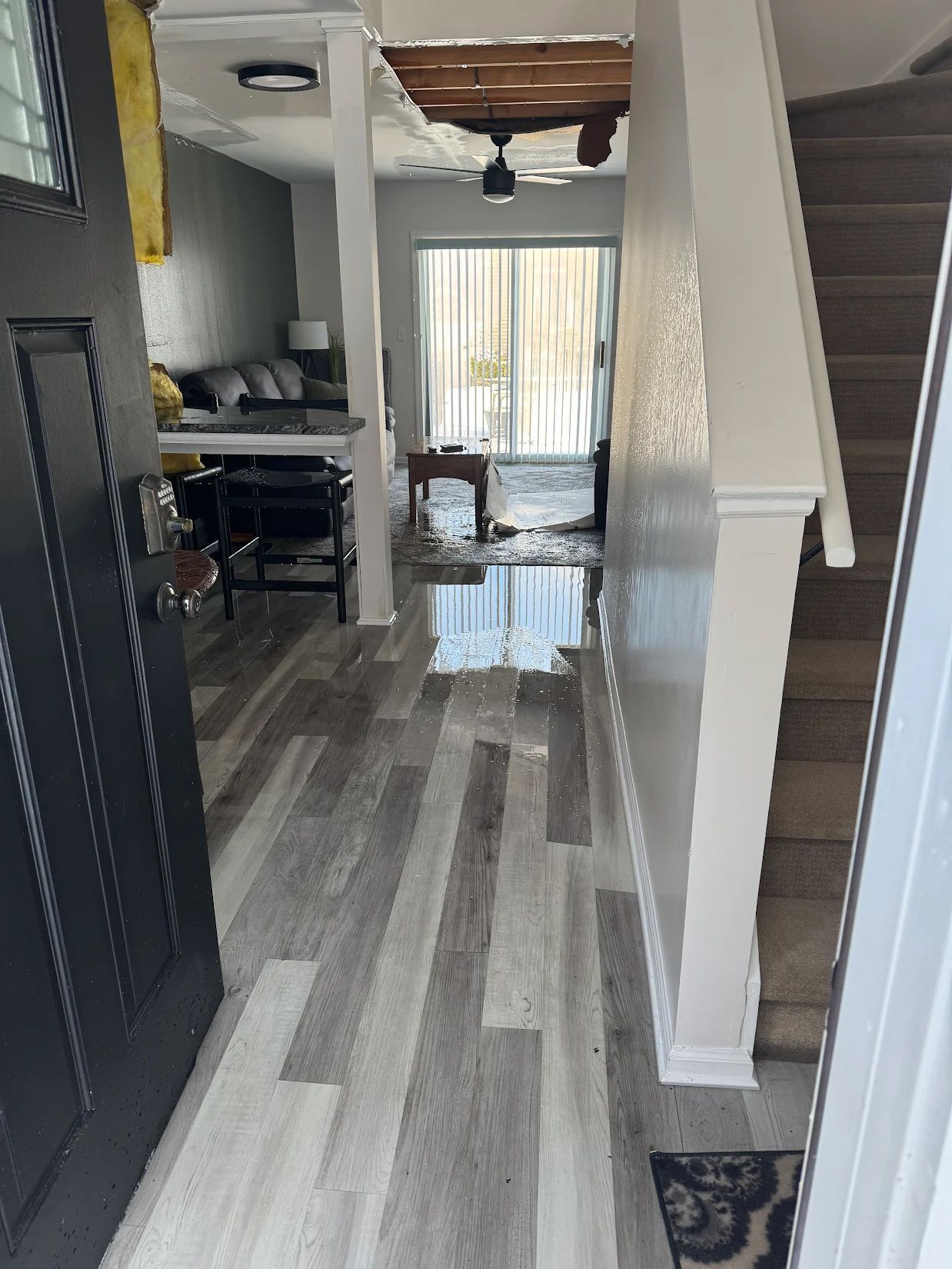Hallway with gray wood flooring, staircase, and open living area with a wet floor near the back window