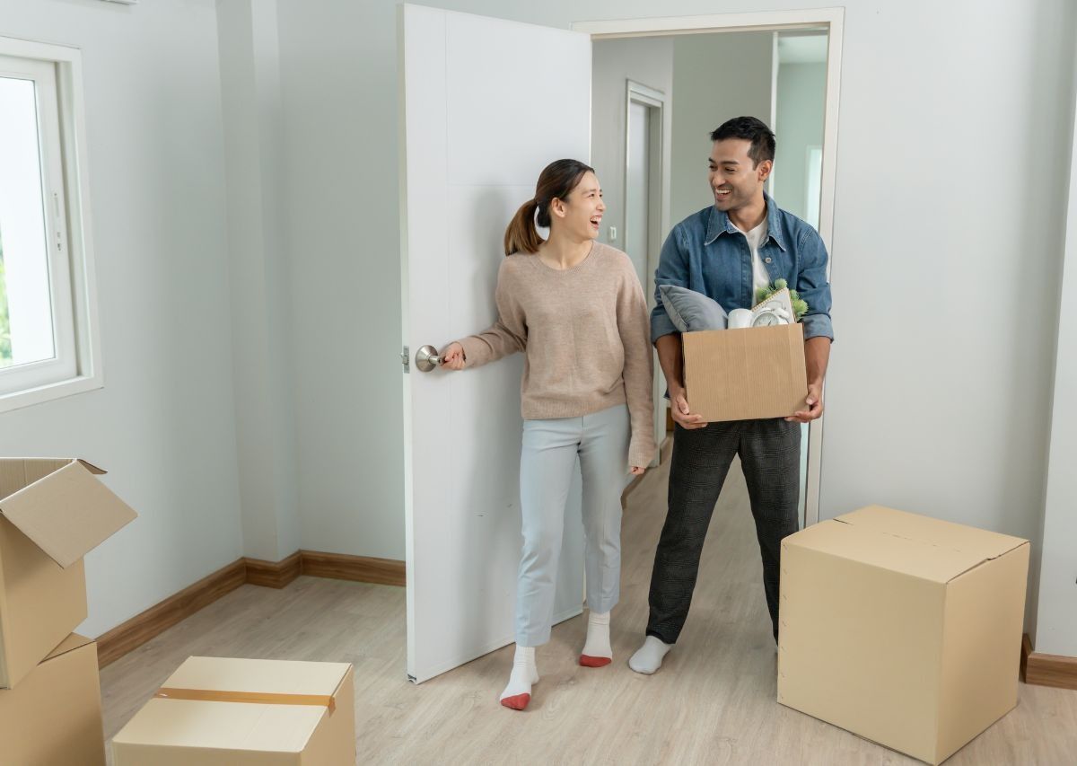 Two people carrying boxes in a bright, mostly empty room, moving into a new home.