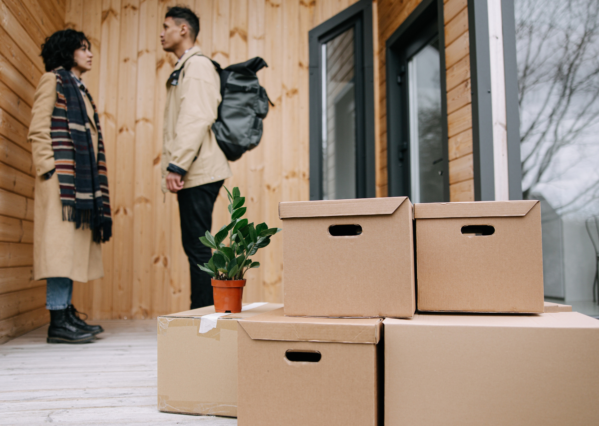 Two people talking beside stacked moving boxes outside a wooden house