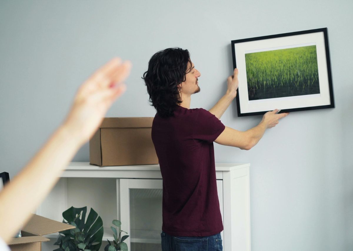 Person hanging a framed landscape photo on a wall in a room with cardboard boxes