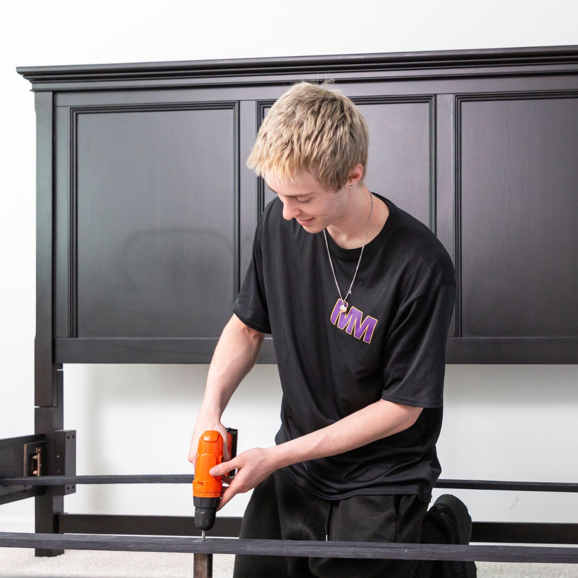 Person in black shirt using an orange drill on a black bed frame indoors