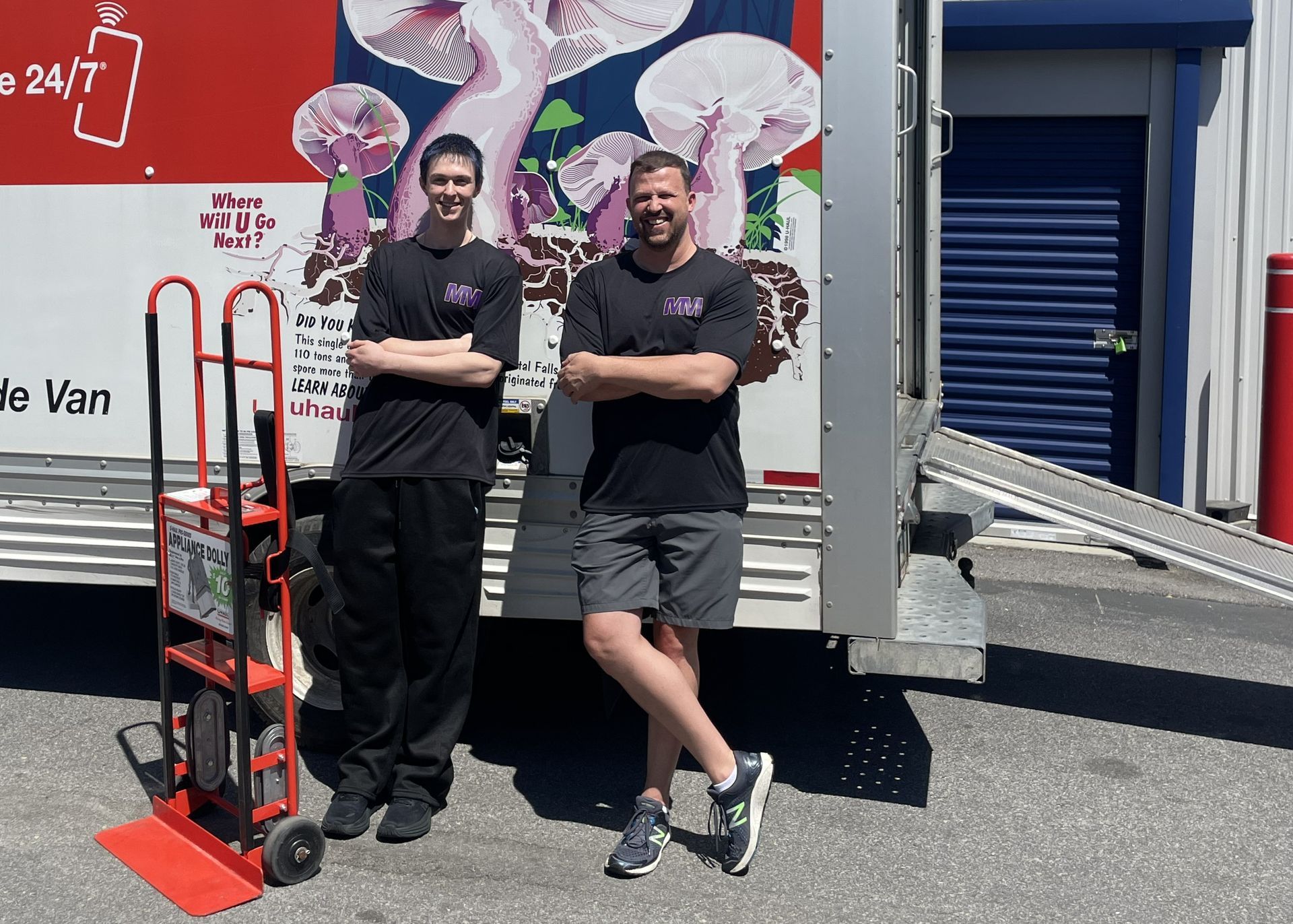 Two people standing beside a red hand truck and a van outside a building, arms crossed, smiling.