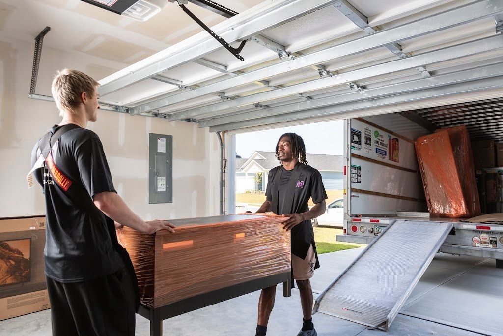 Two people moving a wrapped sofa in a garage with a loading ramp nearby