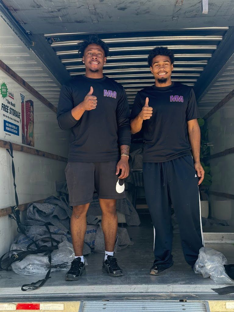 Two smiling workers in black shirts giving thumbs up inside a box truck with stacked bags and tools.
