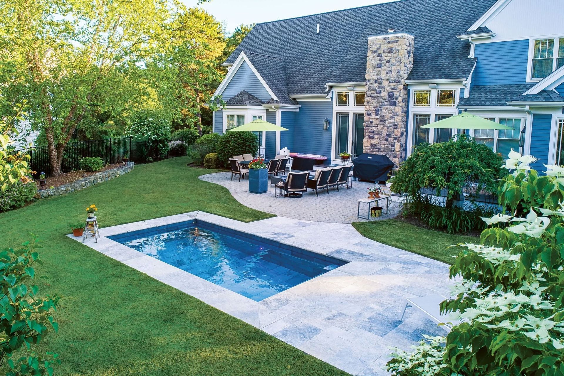 Backyard patio with a blue pool, outdoor seating, and a gray house surrounded by lush greenery