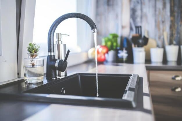 Modern kitchen sink with black faucet, countertop decor, and blurred background plants and utensils