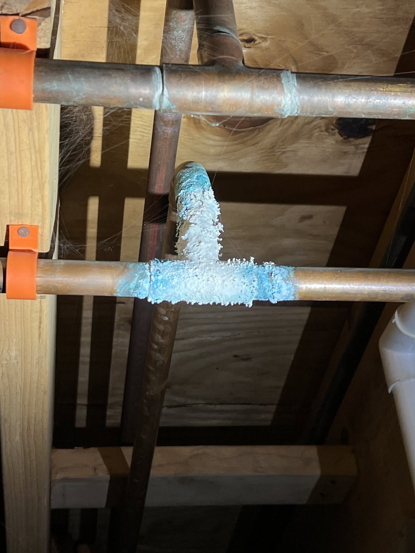 Close-up of corroded metal pipes under a wooden floor, with a blue foamy leak at a joint.