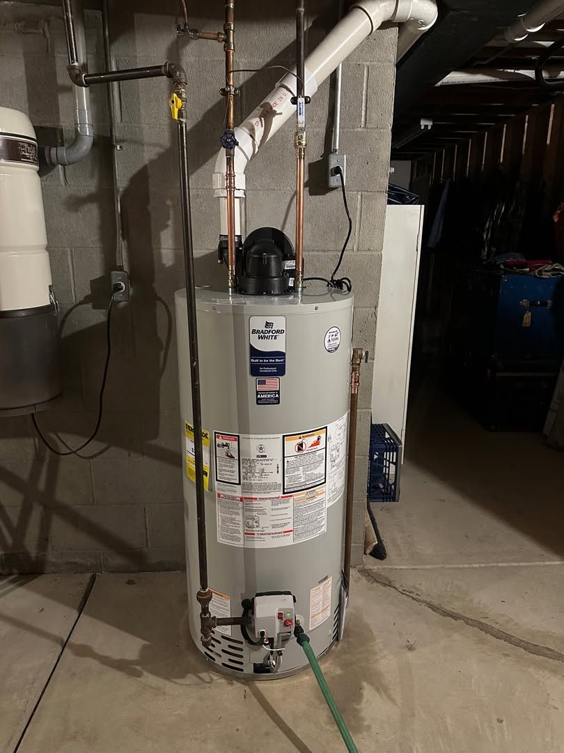 Tall water heater in a dim basement with exposed pipes and a green hose attached.