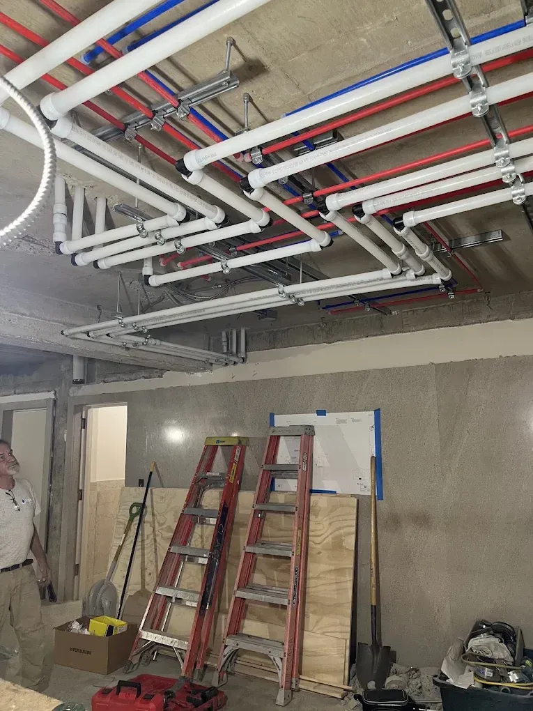 Construction site with exposed ceiling pipes and ladders leaning against a wall in a unfinished room.
