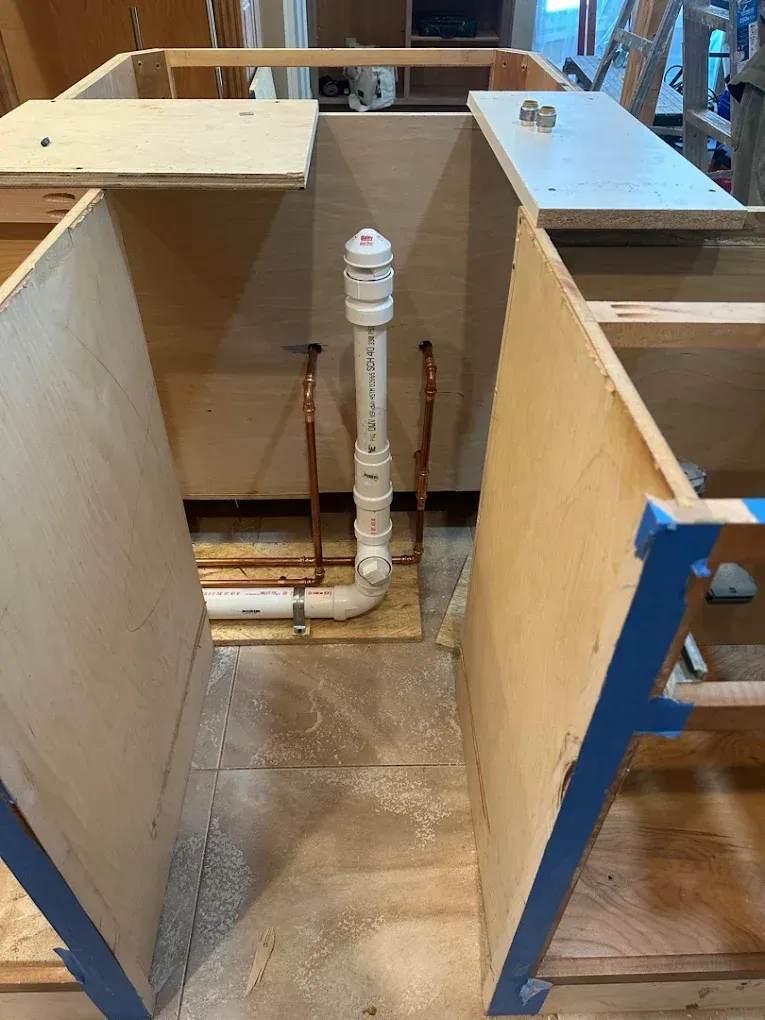 Wood-paneled cabinet interior with exposed white plumbing pipes and a wet floor below.