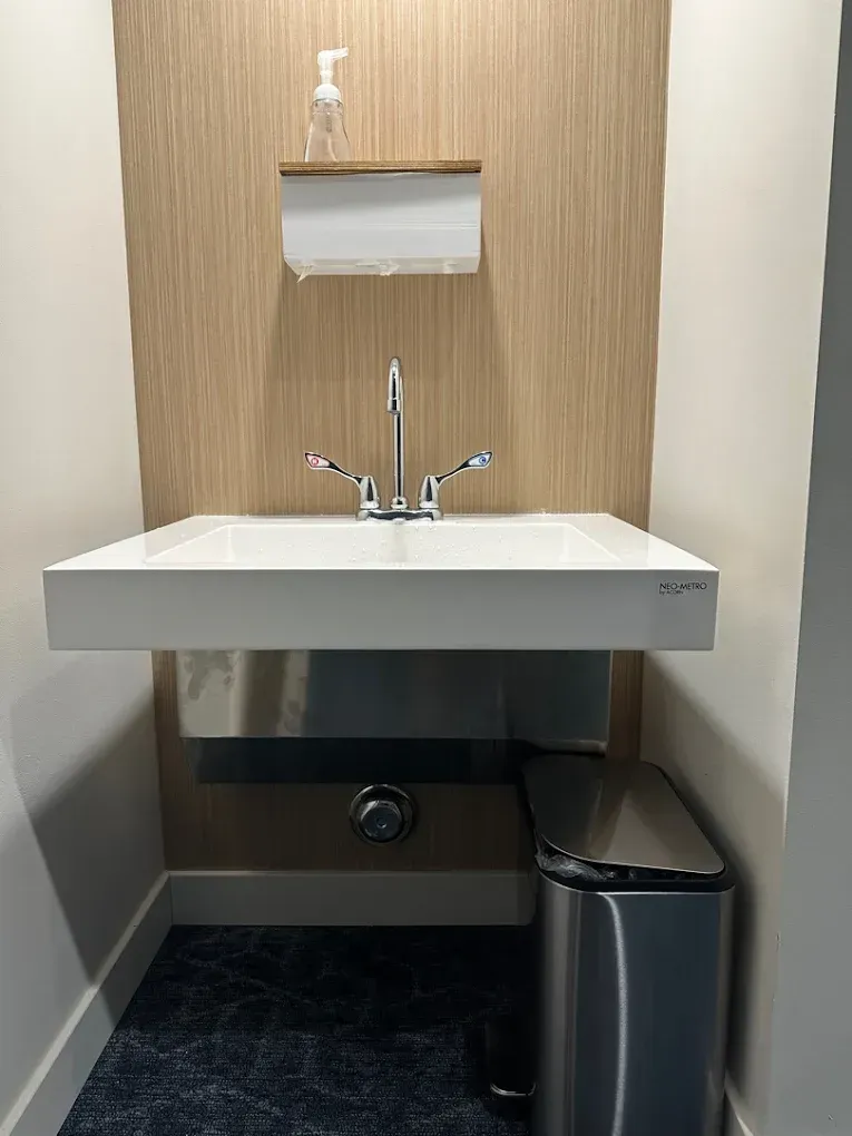 Small white sink in a beige restroom alcove with a chrome faucet and a black trash can below.