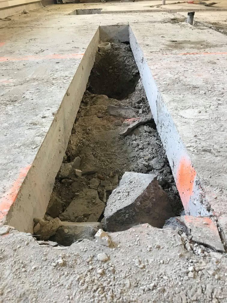 Large rectangular trench cut into a concrete floor with exposed dirt and broken concrete debris.