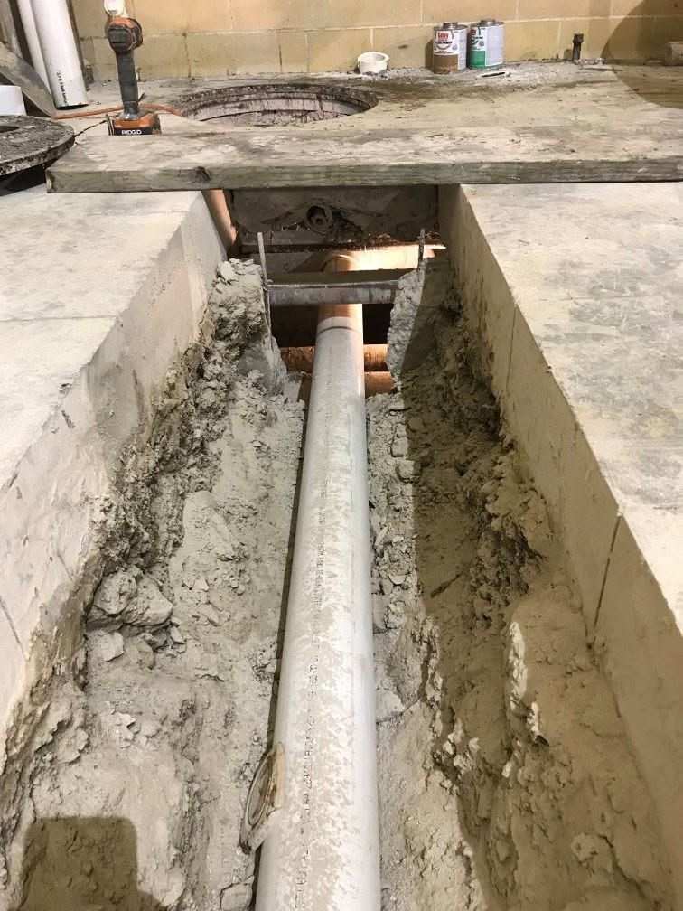 Open trench with exposed white pipe running through a concrete construction site, dirt piled along the sides