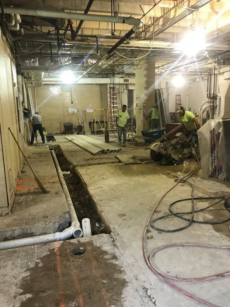 Construction site interior with workers, exposed ceiling, trenches, tools, and scattered cables