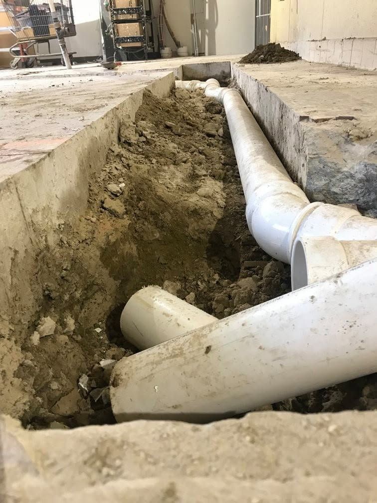 Exposed white PVC pipes in a trench cut through a concrete floor at an indoor construction site