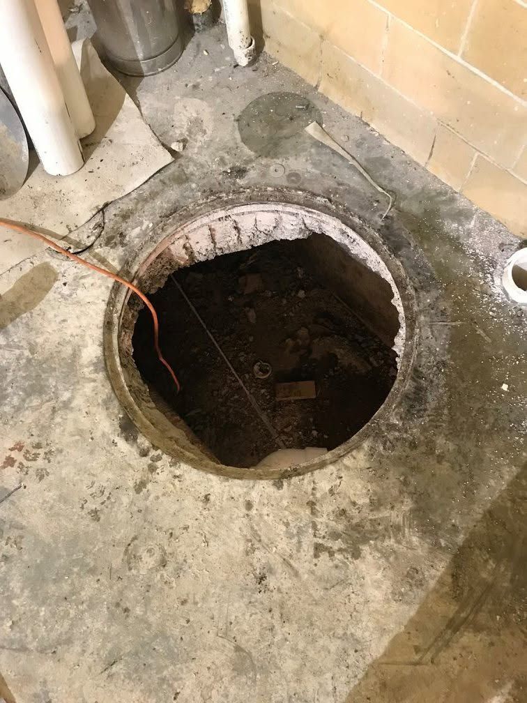 Open floor hatch in concrete room, exposing a dark circular shaft near pipes and a brick wall.
