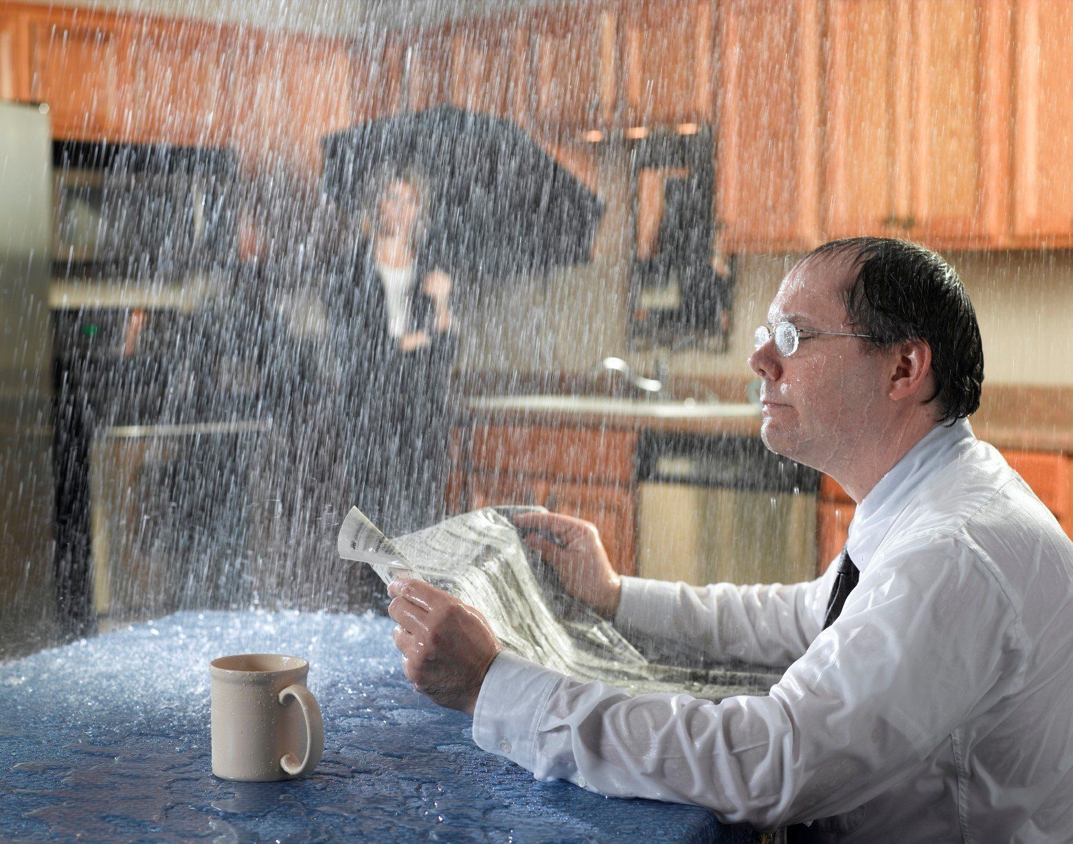 Man in white shirt reading beside a kitchen table as rain pours through the ceiling onto him.
