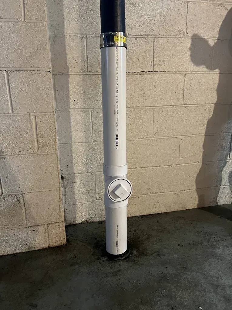 White vertical pipe with a circular access panel mounted against a concrete block wall.