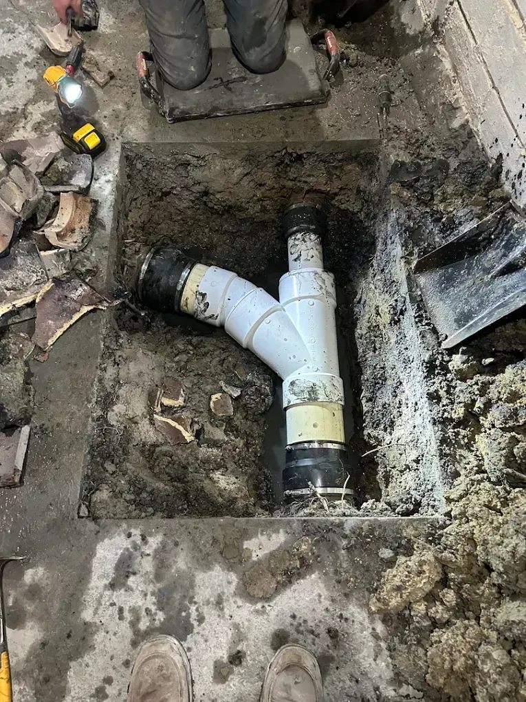 Open trench with white PVC plumbing pipes and muddy soil, with gloved hands working nearby.