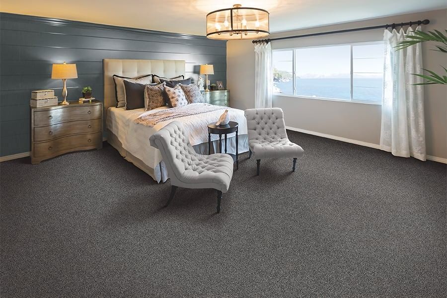 Bedroom with gray carpet, bed, and chairs near a large window overlooking the ocean.