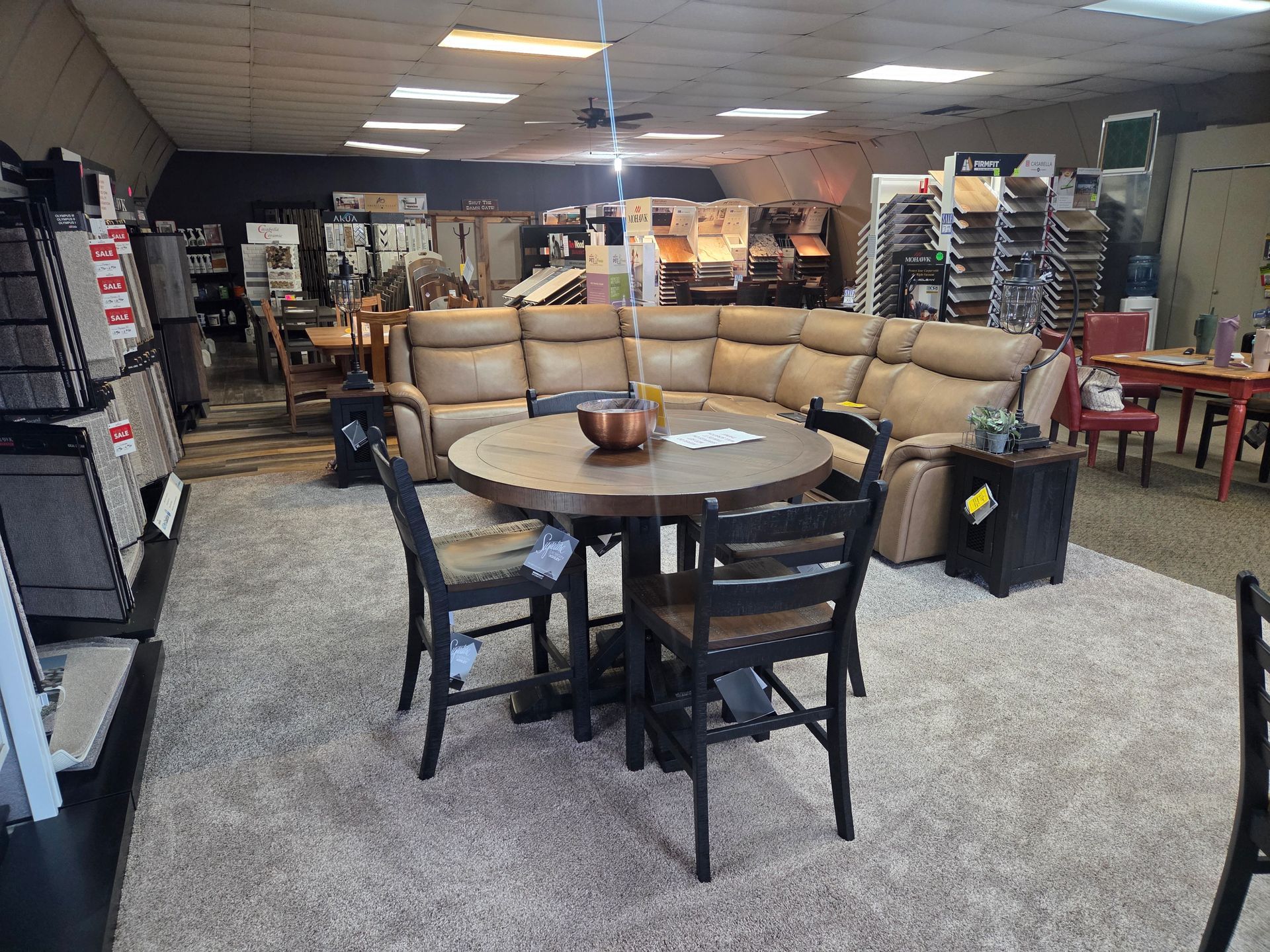 Furniture store interior featuring a sectional sofa, dining set, and displays of flooring samples.