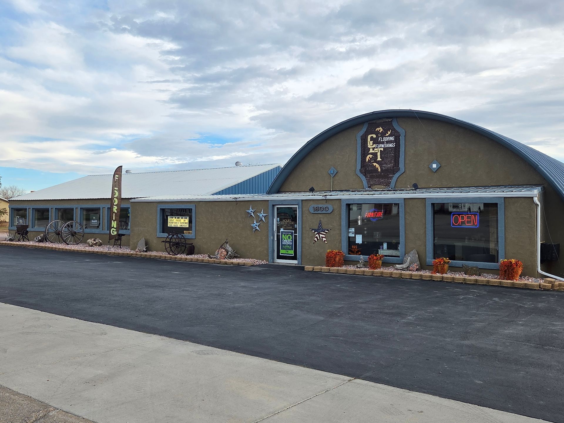 Tan commercial building with arched roof, windows, and decorative emblem above entrance. Paved parking area in front.