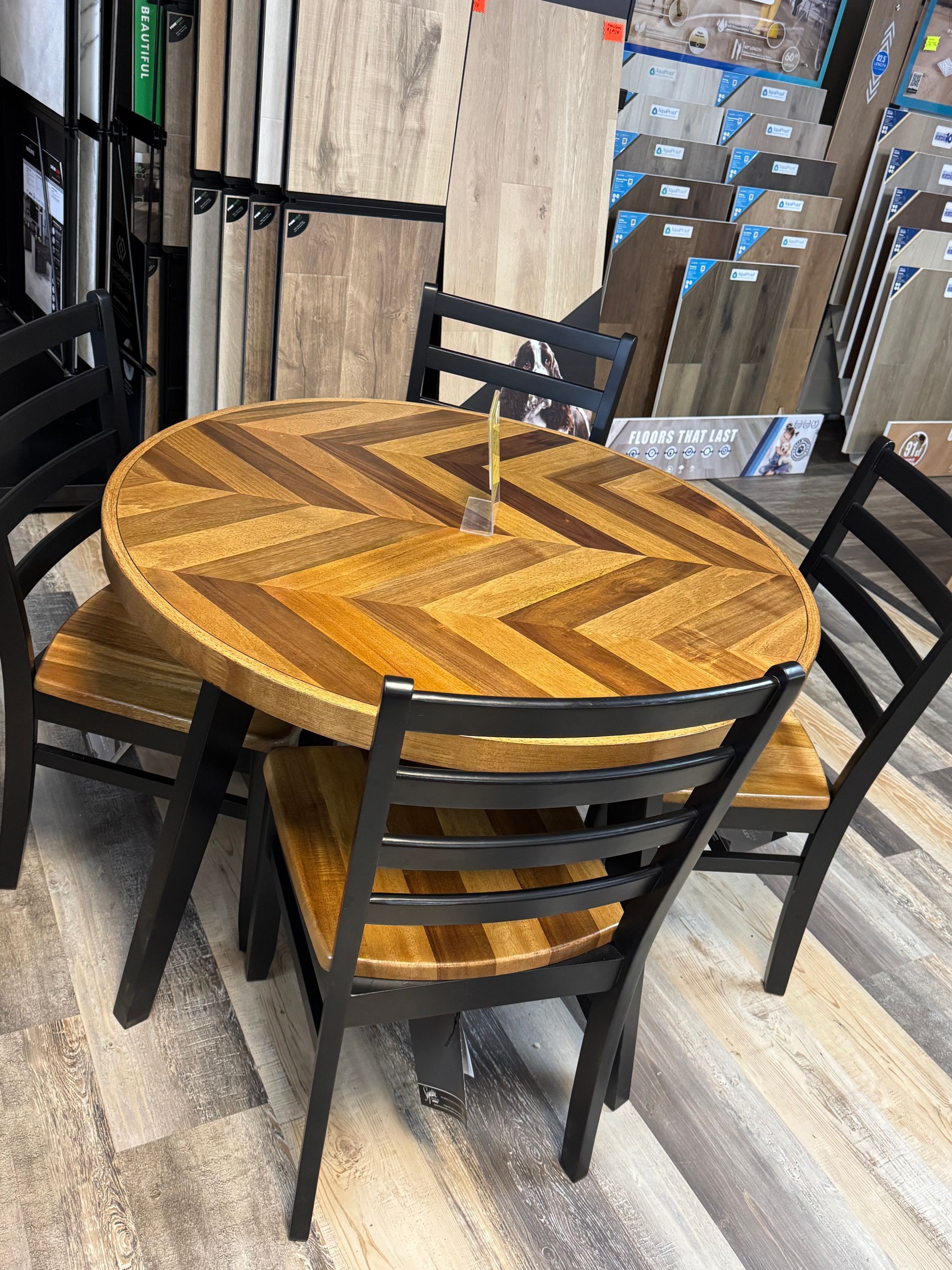 Round wooden table with herringbone pattern and four black chairs in a showroom setting.