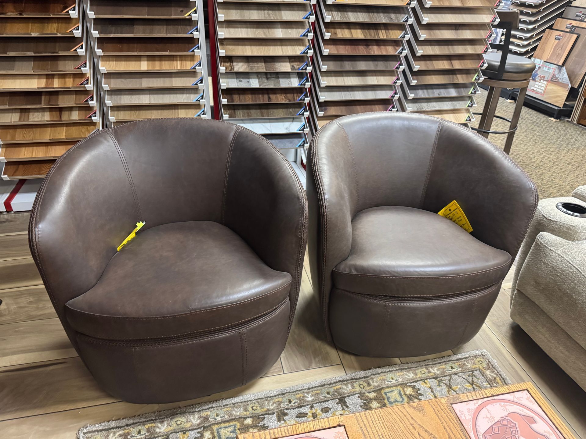 Two brown leather barrel chairs in a showroom, with wood flooring samples in the background.