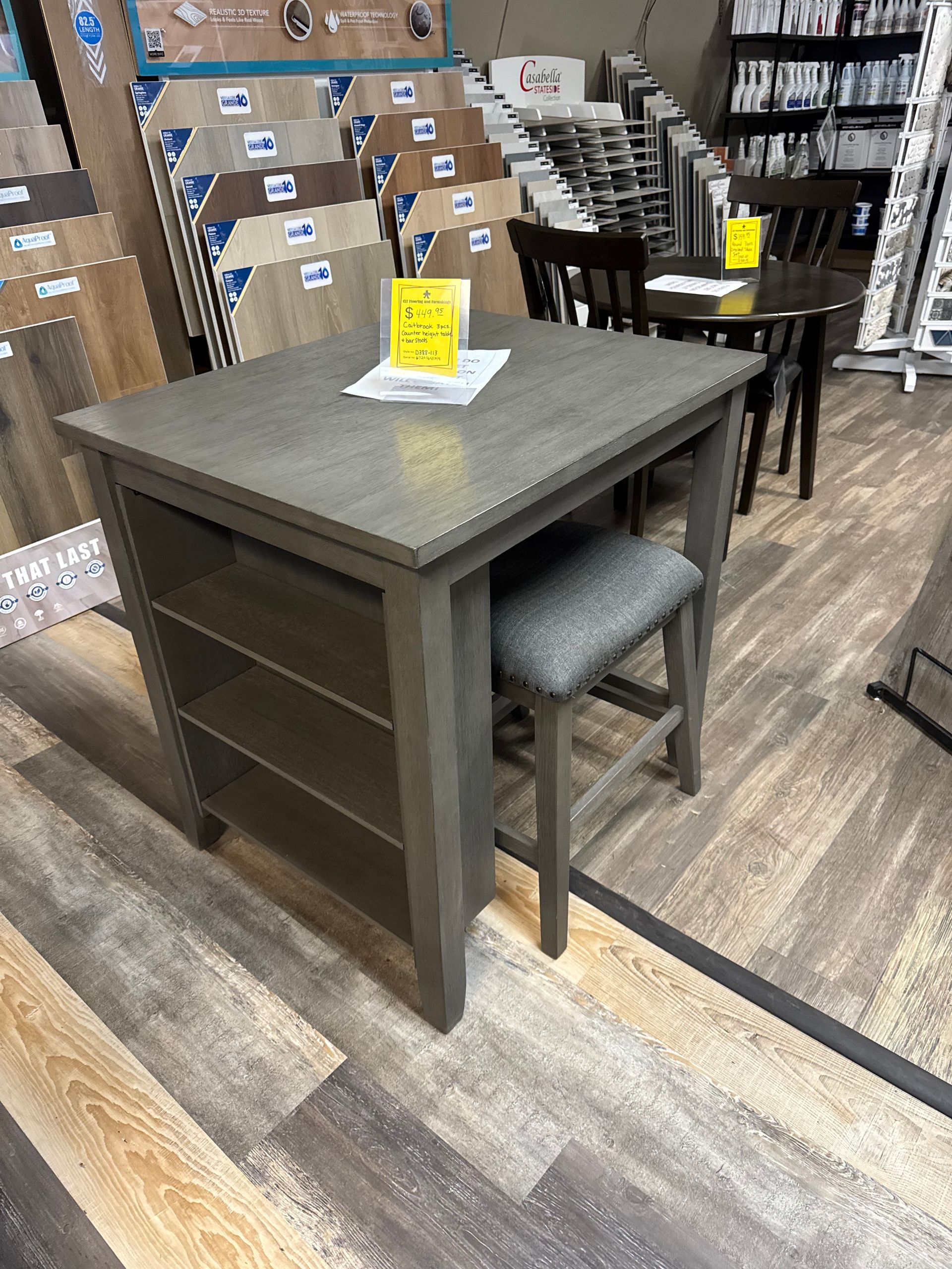 Gray square dining table with built-in shelving, a gray upholstered stool, and wood flooring in a store.