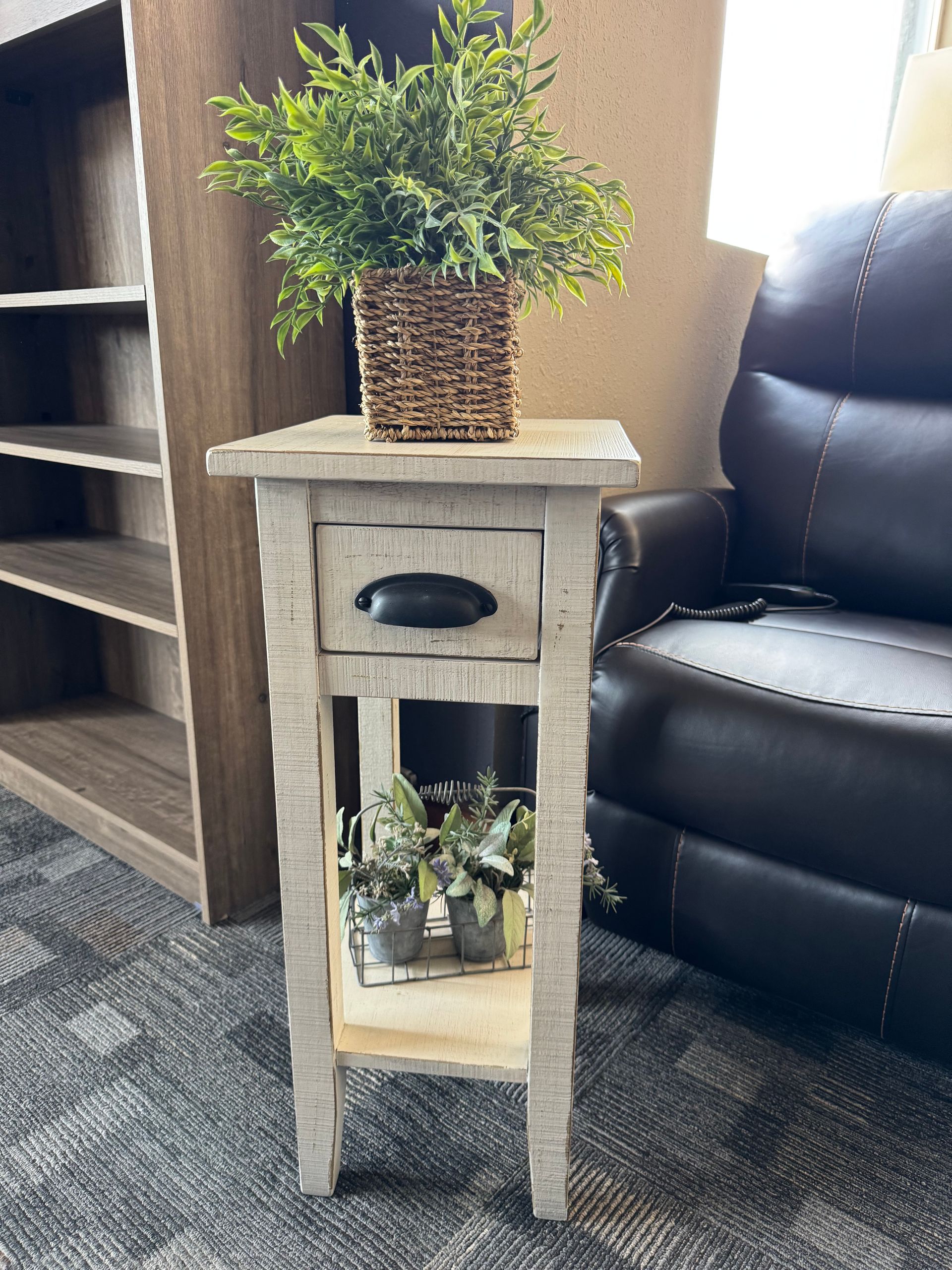 Small, distressed white side table with plant in basket, drawer, and shelf. A recliner is in background.