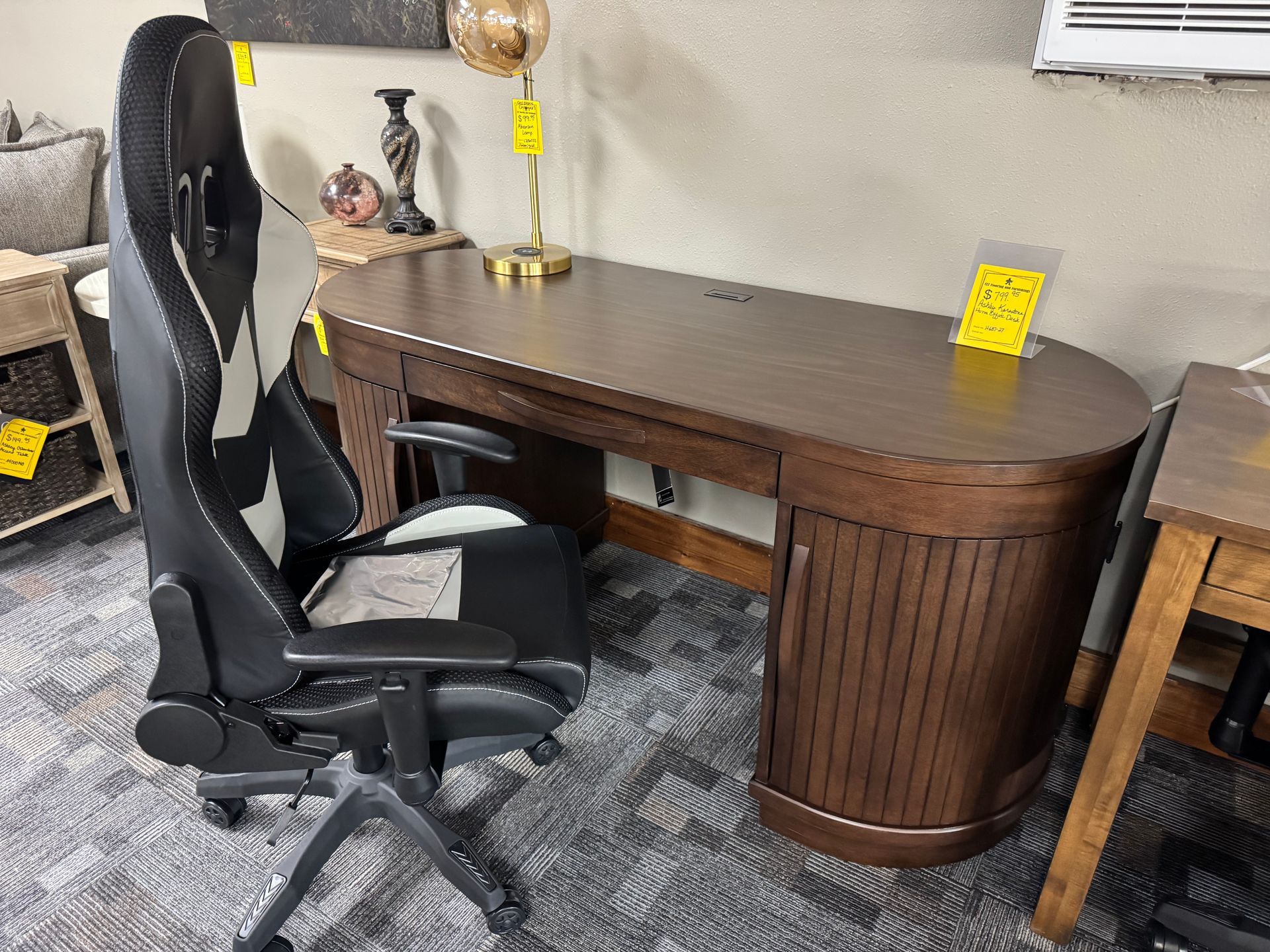 A dark wood desk with a gamer chair and a lamp in a store setting.