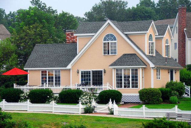 A light peach two-story house with a white picket fence, green bushes, and a red patio umbrella.