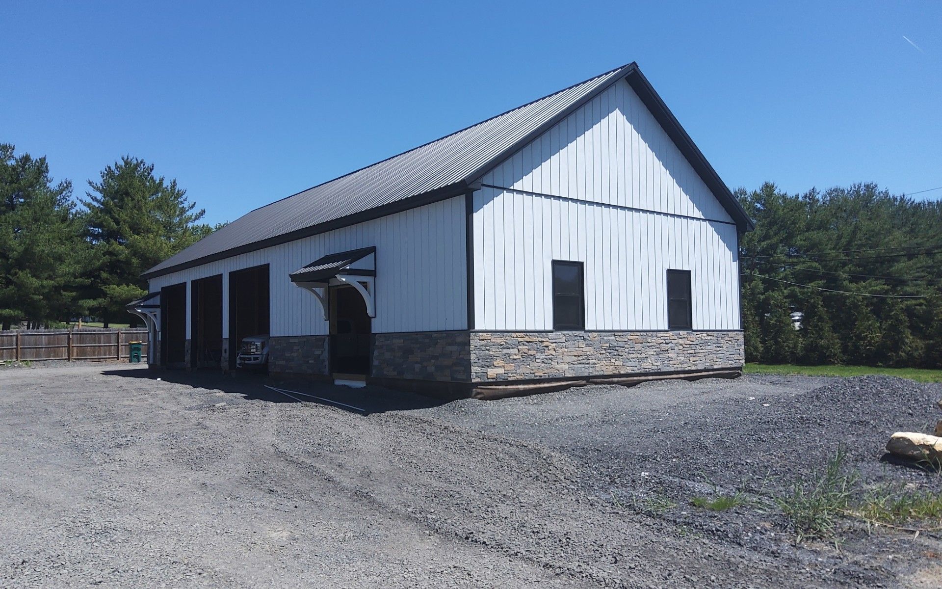White barn-style building with stone foundation and black doors on a gravel lot under a blue sky