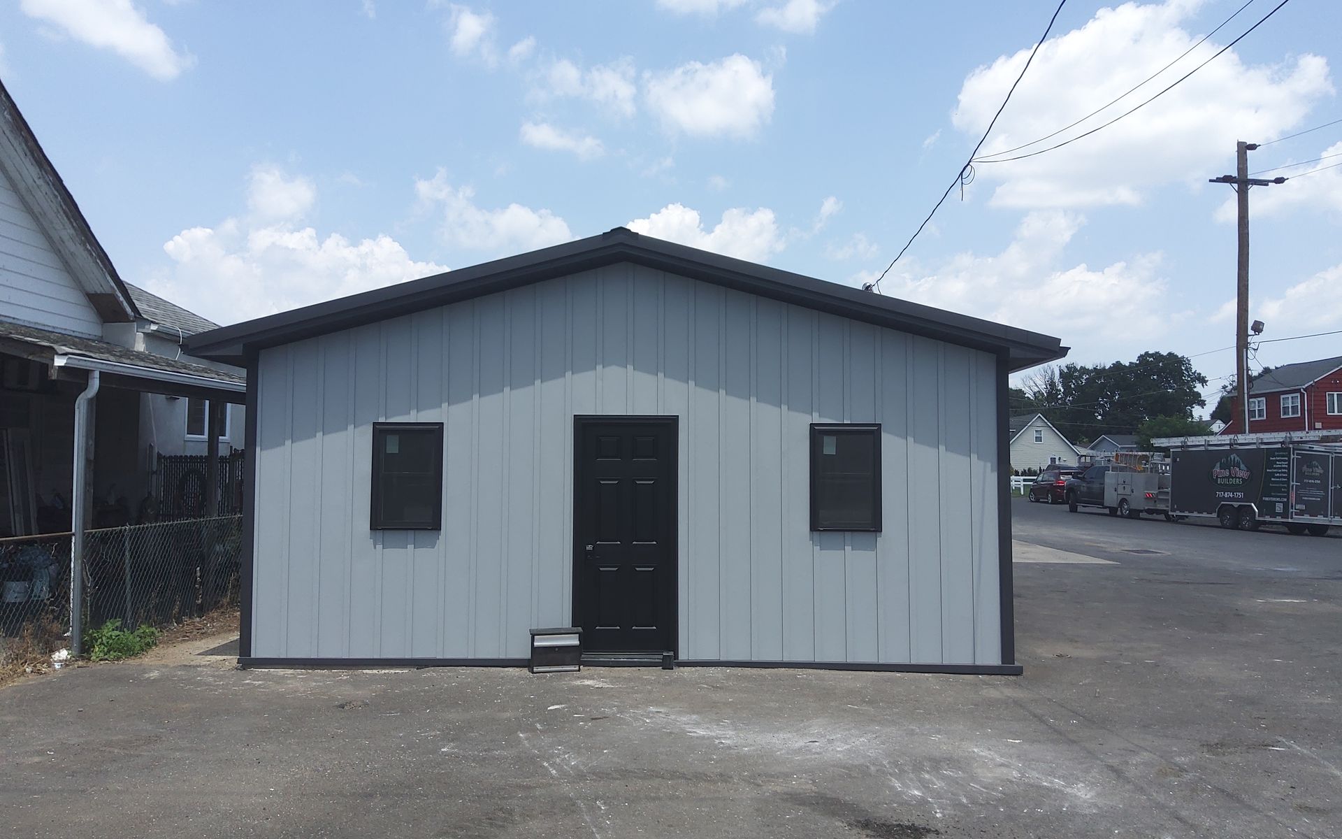 Small gray building with a dark door and windows on a paved lot under a partly cloudy sky