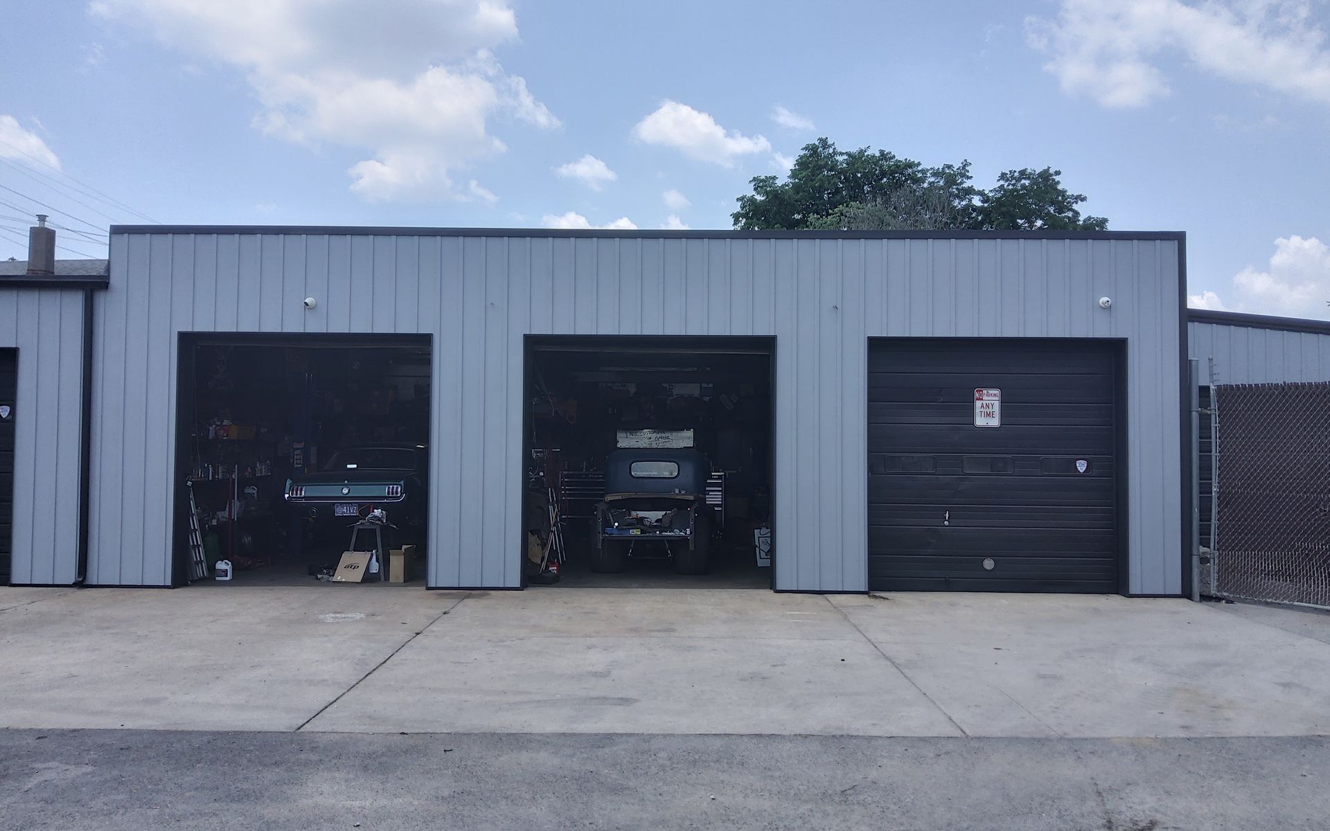 Gray industrial garage with three black roll-up doors on a concrete lot under a cloudy sky