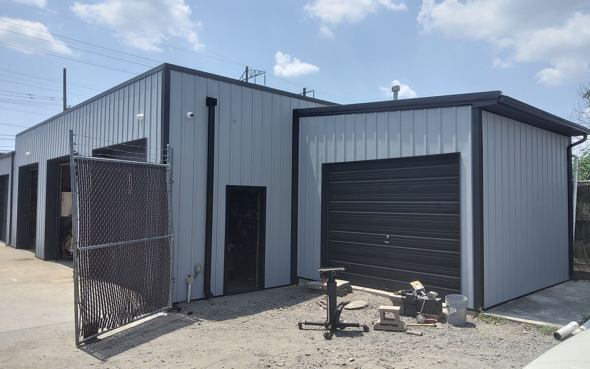 Gray industrial metal building with open doors and a black wrought-iron gate outside under a partly cloudy sky