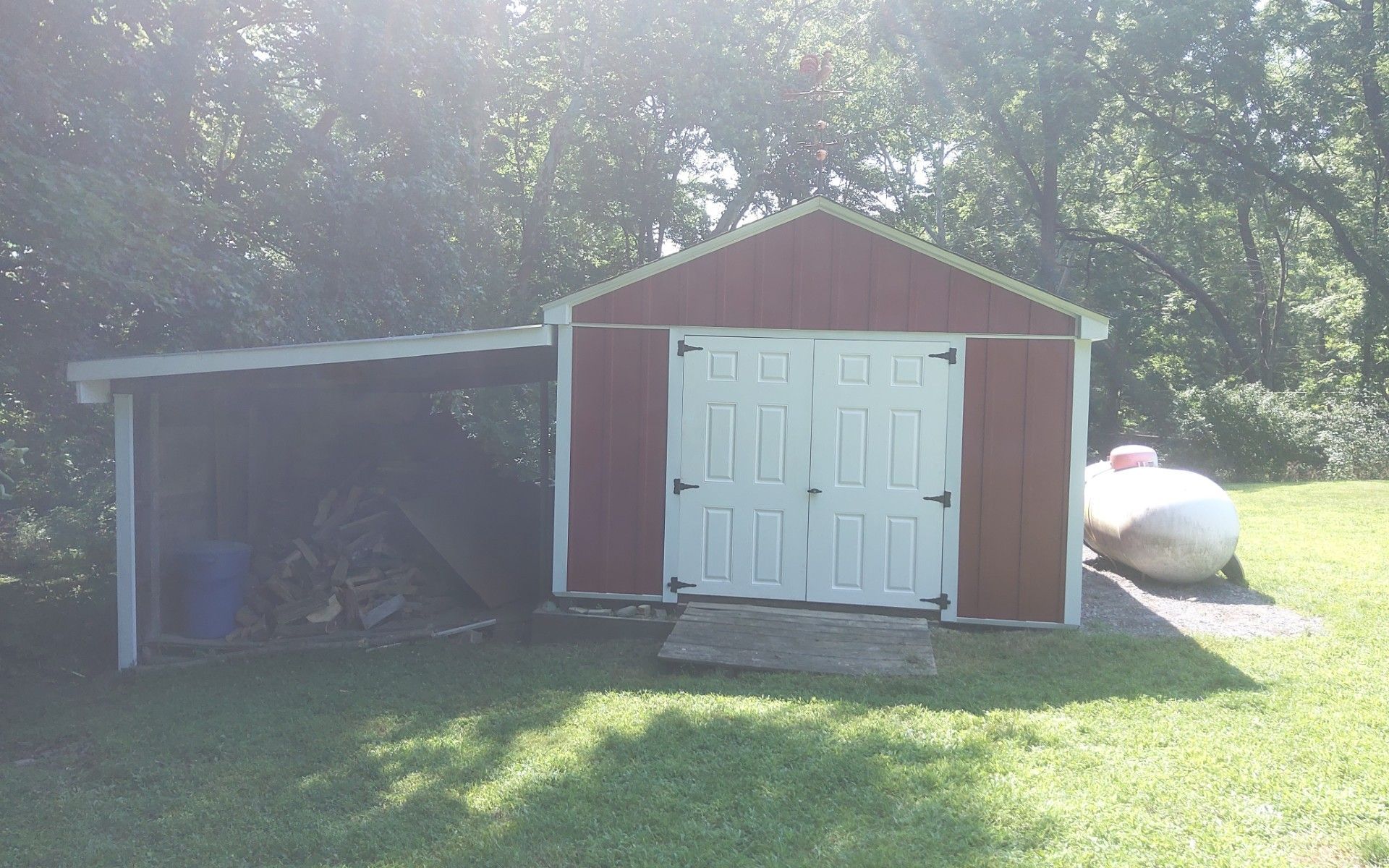 Red barn-style shed with white double doors and a small open lean-to, surrounded by trees and grass.