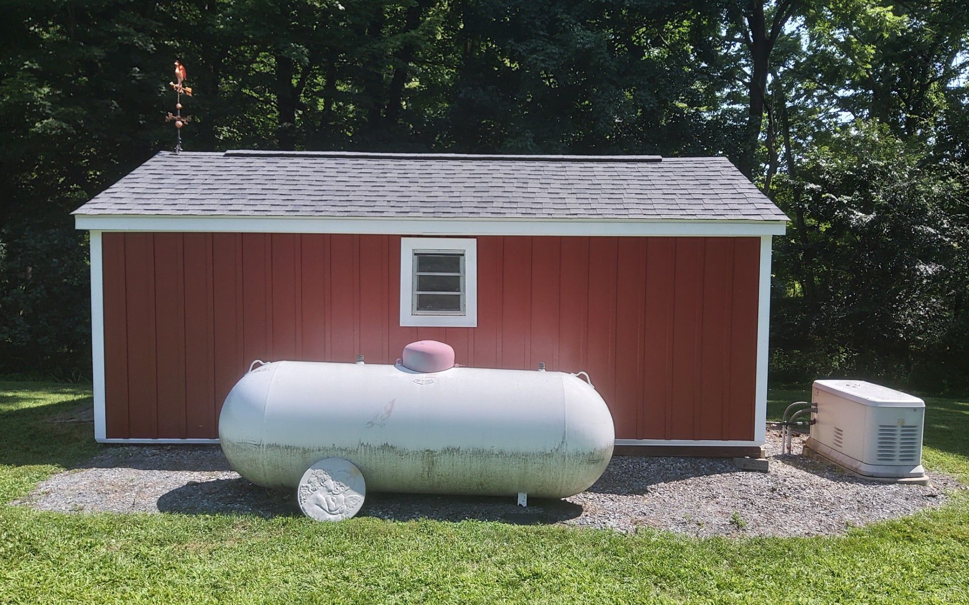 Red shed with a white propane tank and gravel yard in front, surrounded by trees.