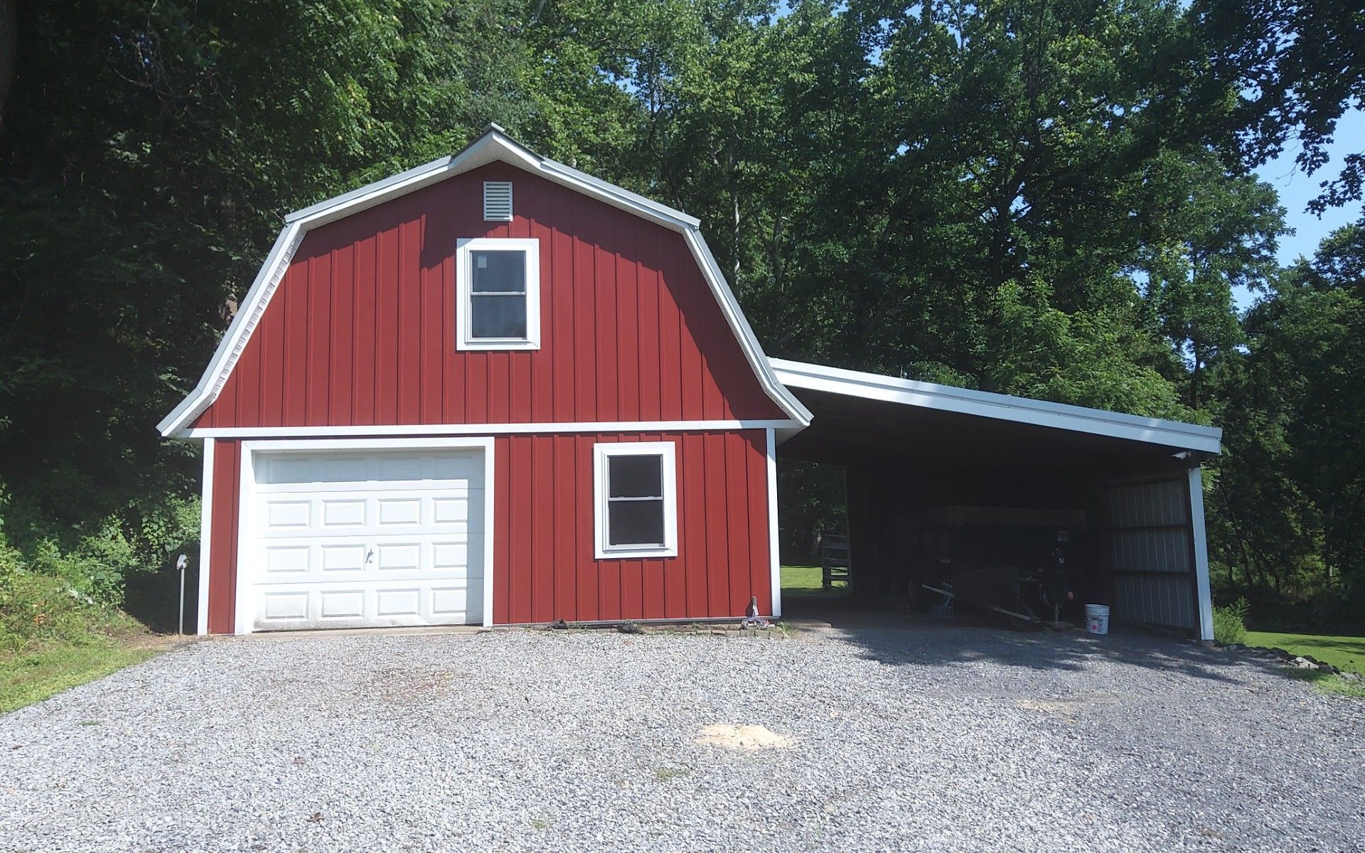 Red barn with white trim, garage door, and attached open carport beside a gravel driveway and trees.